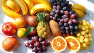 Colorful assortment of fresh tropical fruits including mangoes, bananas, and grapes arranged on a white marble surface with natural sunlight, vibrant and appetizing presentation