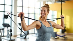 Woman in bright fitness studio performing strength training with resistance bands, smiling, natural daylight streaming through large windows, modern equipment visible