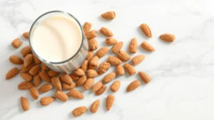Overhead view of a clear glass filled with creamy unsweetened almond milk beside fresh whole almonds and almond pieces scattered on a white marble surface, natural daylight, photorealistic, wellness aesthetic